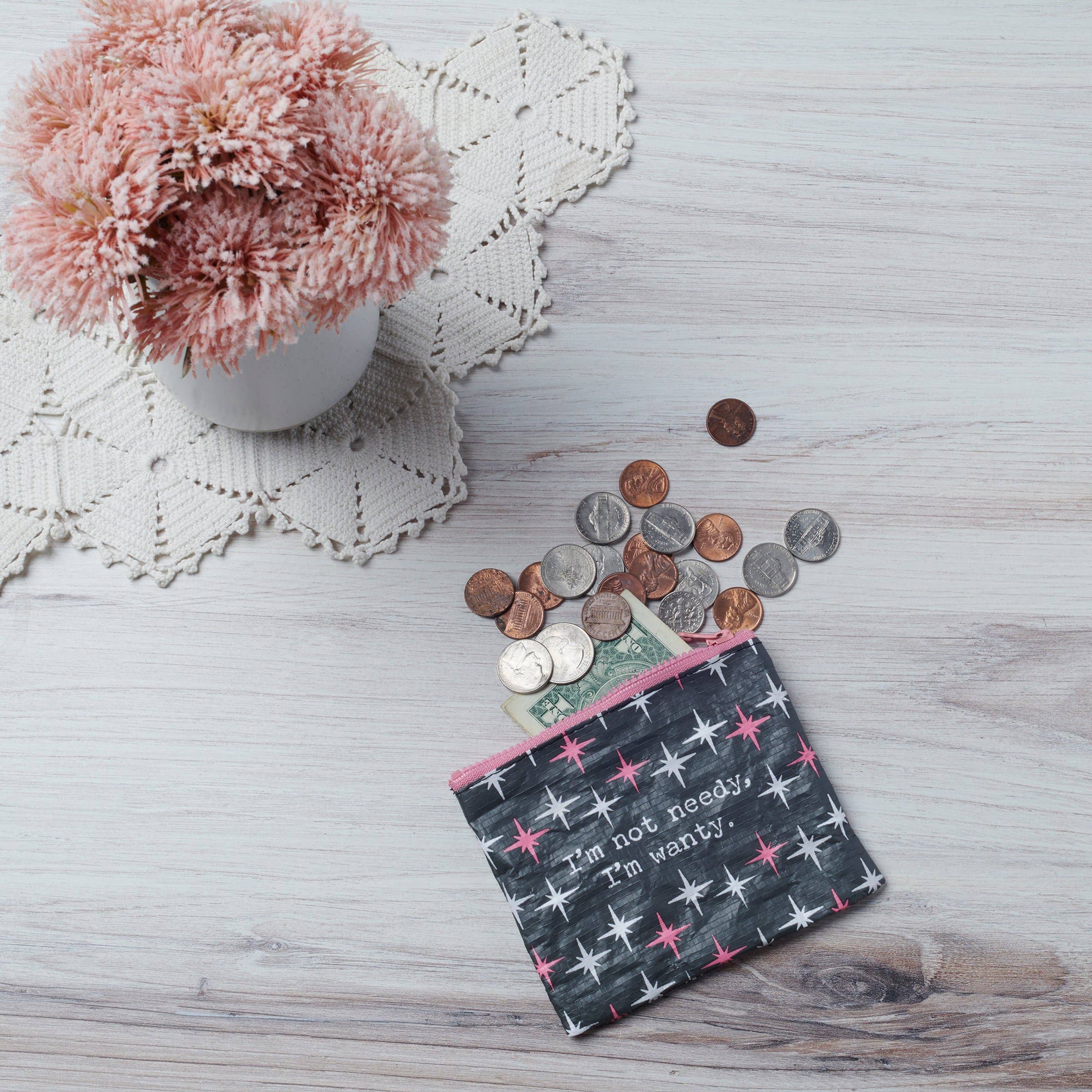 A patterned pouch with the text "I'm not needy, I'm wanty" spills coins and bills on a light wooden table. Nearby, pink flowers sit in a vase on a lace mat.