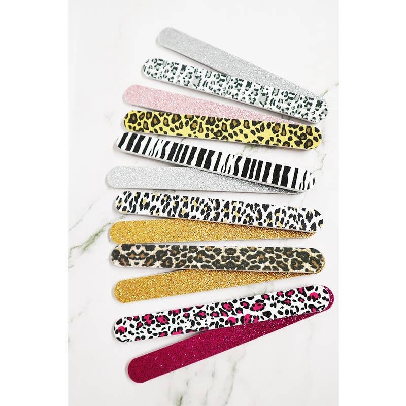 An assortment of ten glittery and patterned emery board nail files fanned out on a white marble background, featuring zebra stripes, cheetah print, and hot pink leopard print.