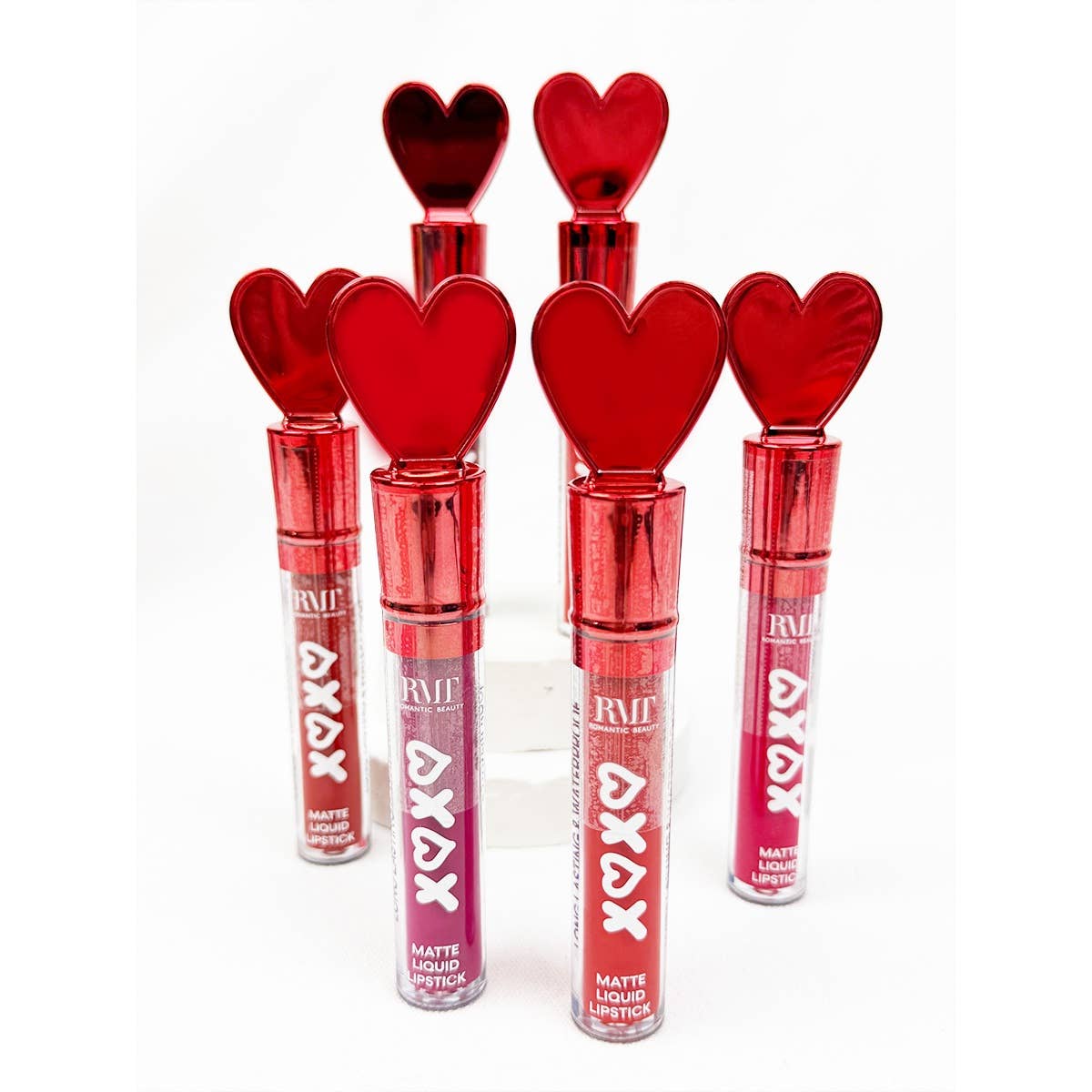 Array of six matte liquid lipsticks with red heart-shaped caps, standing upright. Varying shades suggest a playful, romantic theme.
