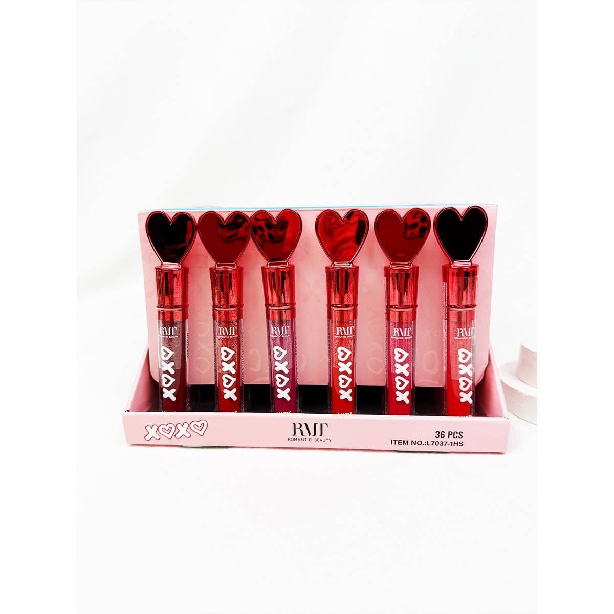 Display tray of heart-shaped red and black lip gloss tubes labeled “XOXO,” arranged upright in pink packaging from Romantic Beauty, with six glosses visible in varying red shades.