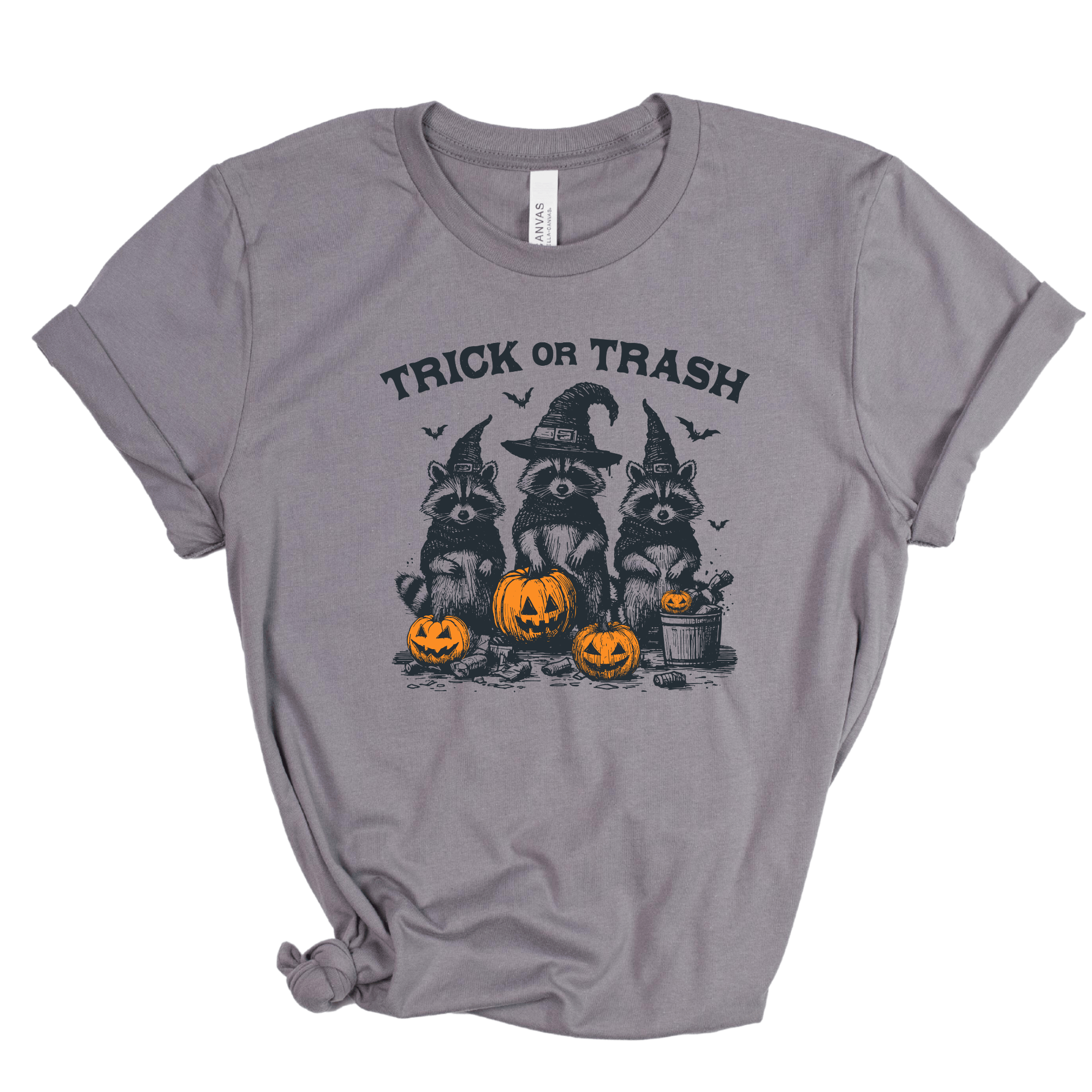 A heather gray short-sleeve t-shirt with a distressed graphic of three raccoons dressed as witches wearing pointed hats, standing behind three jack-o'-lantern pumpkins. The text 'TRICK OR TRASH' is printed above the graphic.