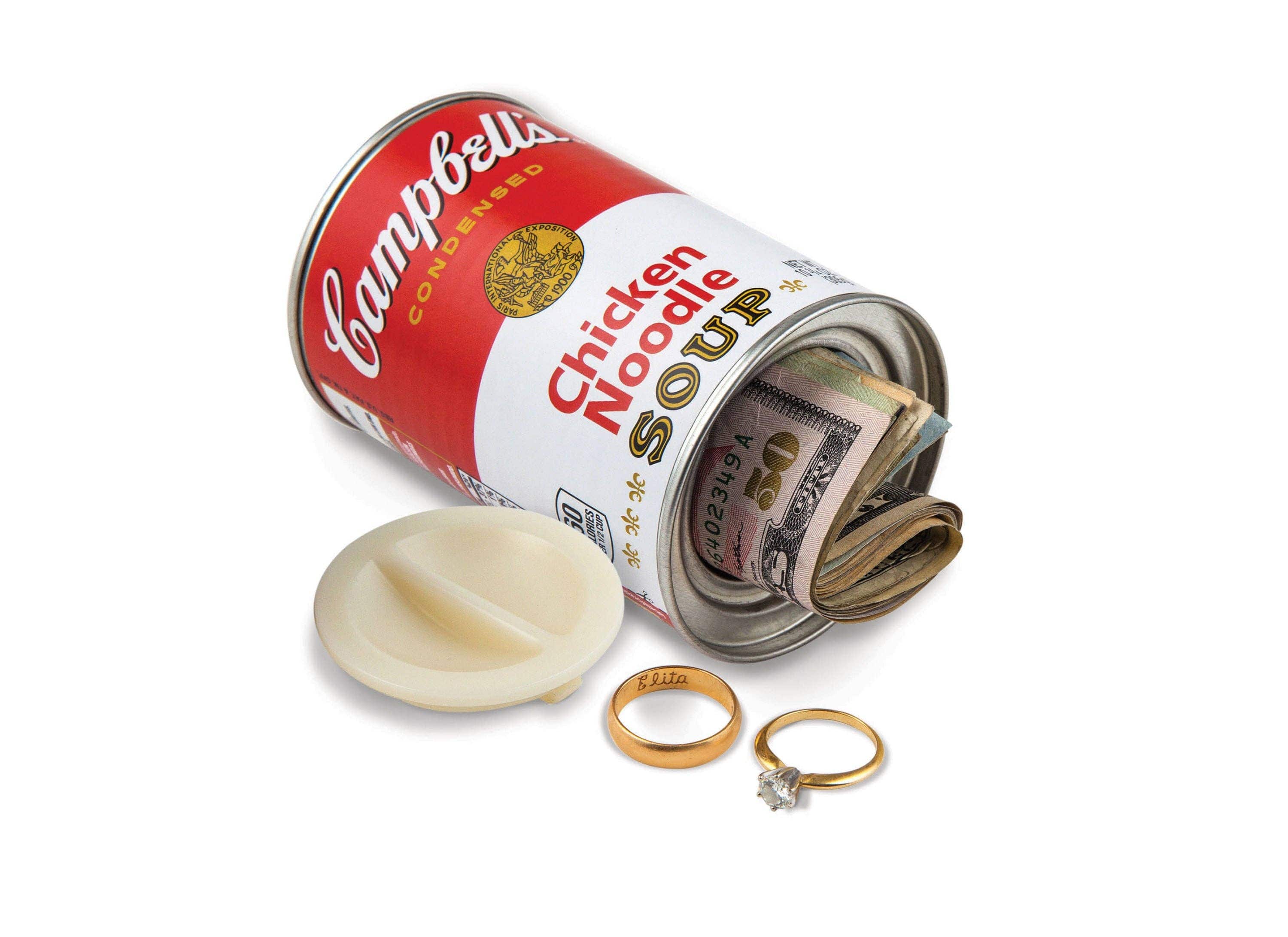 A Campbell's Chicken Noodle Soup can, with its lid removed, reveals rolled U.S. dollar bills inside. Two rings are placed nearby, evoking a sense of hidden treasure.