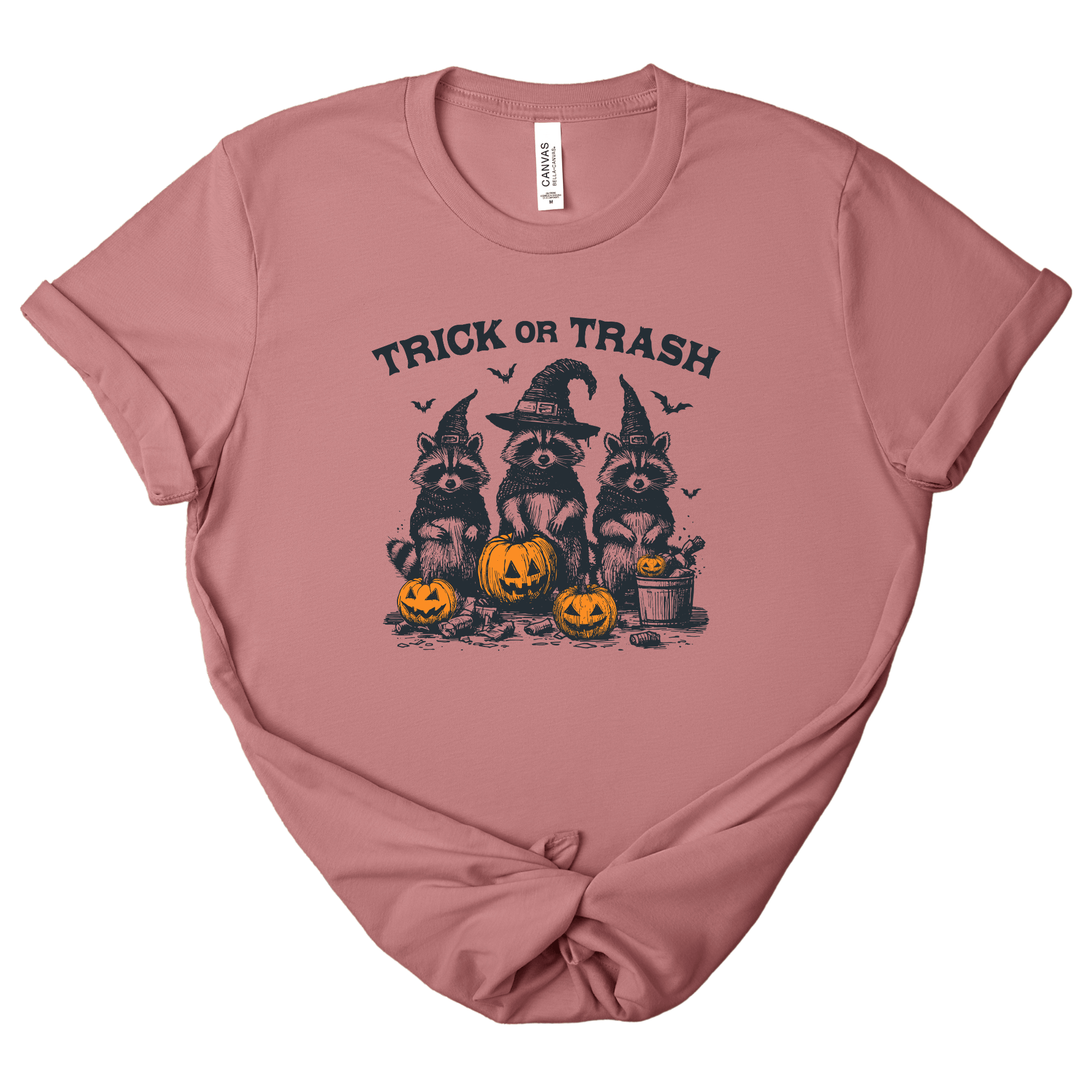 A dusty rose short-sleeve t-shirt with a 'Trick or Trash' Halloween graphic featuring three raccoons in witch hats and jack-o'-lanterns, laid flat on a white background.