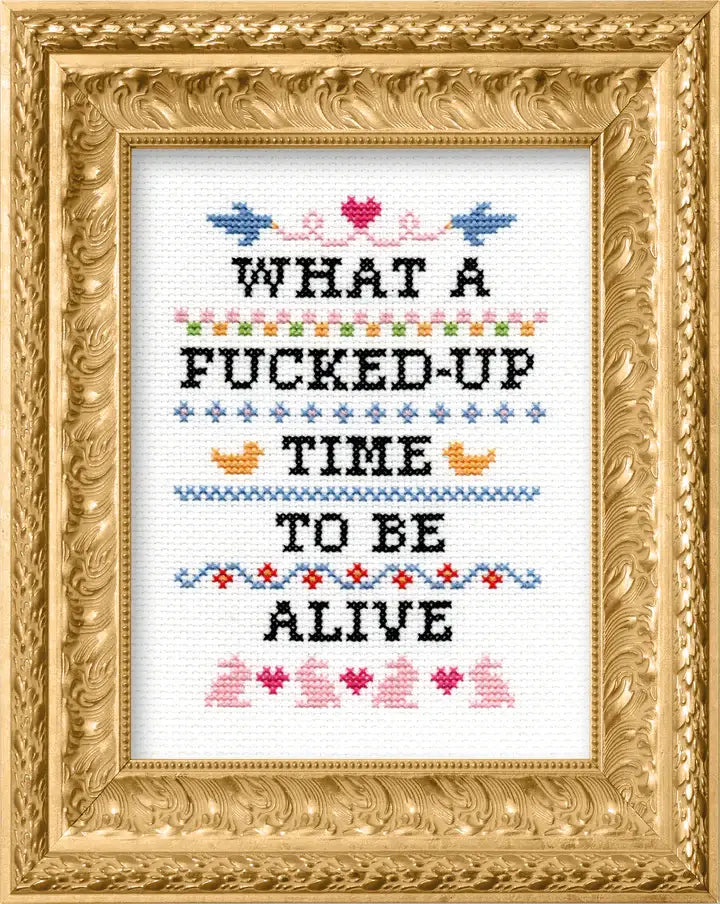 Cross-stitched text in a decorative gold frame "What a fucked up time to be alive."