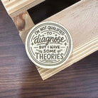 Circular sticker with text on a wooden surface.Text states "I'm not qualified to diagnose, but I have some theories."