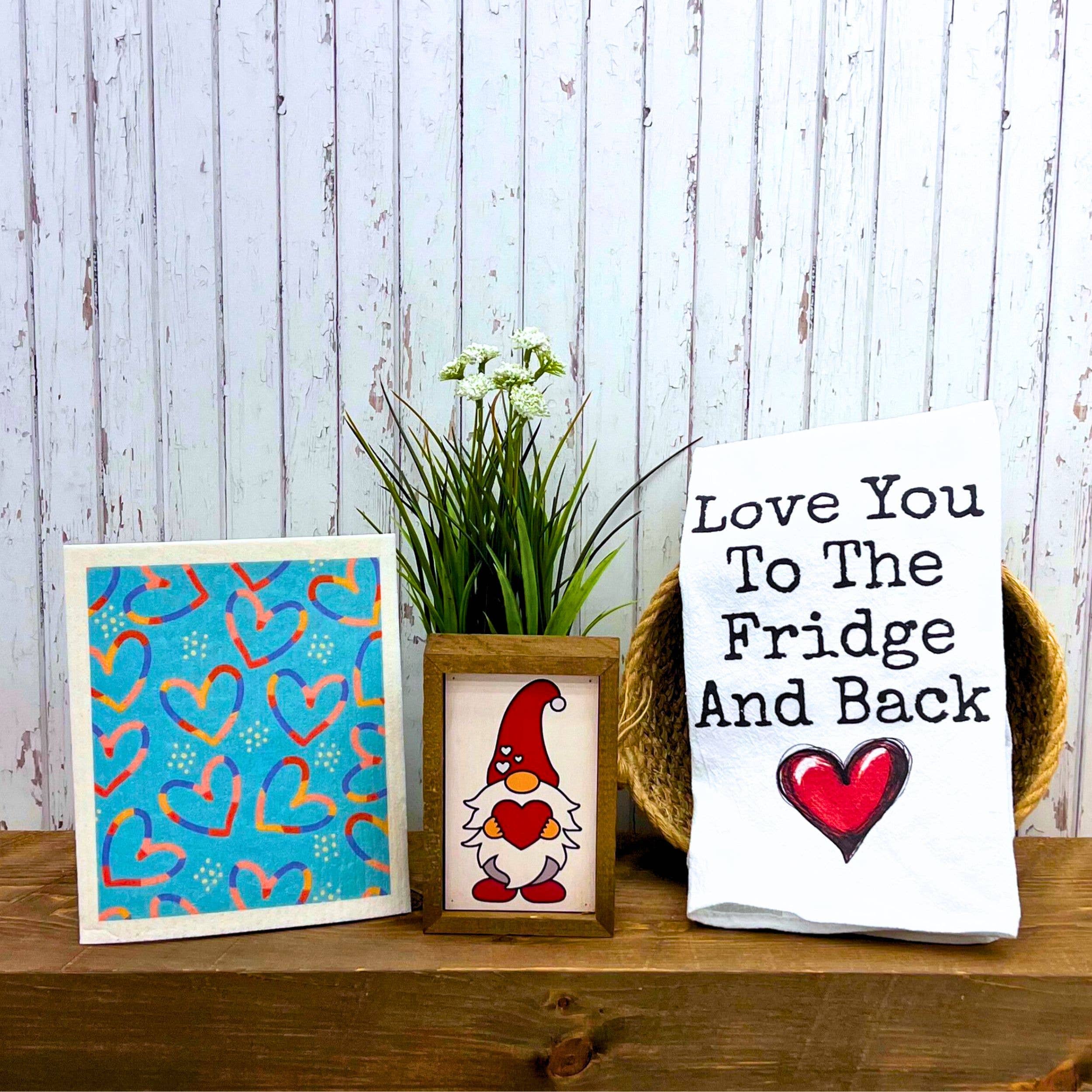 Decorative setup featuring a colorful heart pattern on a blue background, a framed gnome with a heart, and a towel stating “Love You To The Fridge And Back,” evoking warmth.