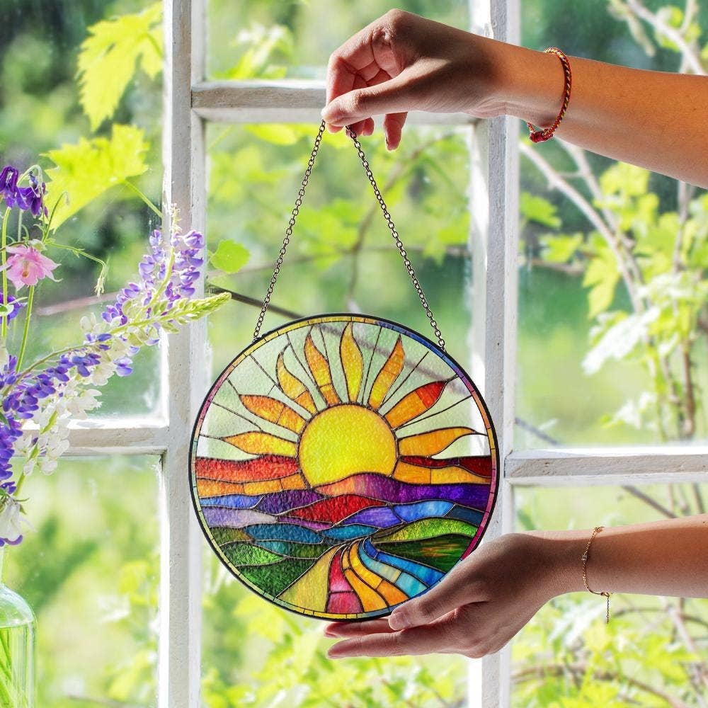 Colorful sun-shaped stained glass panel held by a hand in front of a window with greenery.