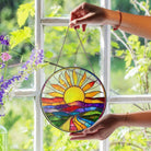 Colorful sun-shaped stained glass panel held by a hand in front of a window with greenery.