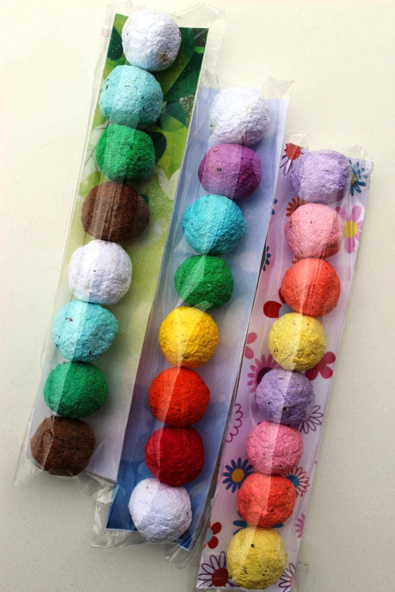 Three packs of colorful seed balls in transparent plastic. Each pack has balls lined in vibrant hues, such as green, blue, pink, and brown, with floral backgrounds.