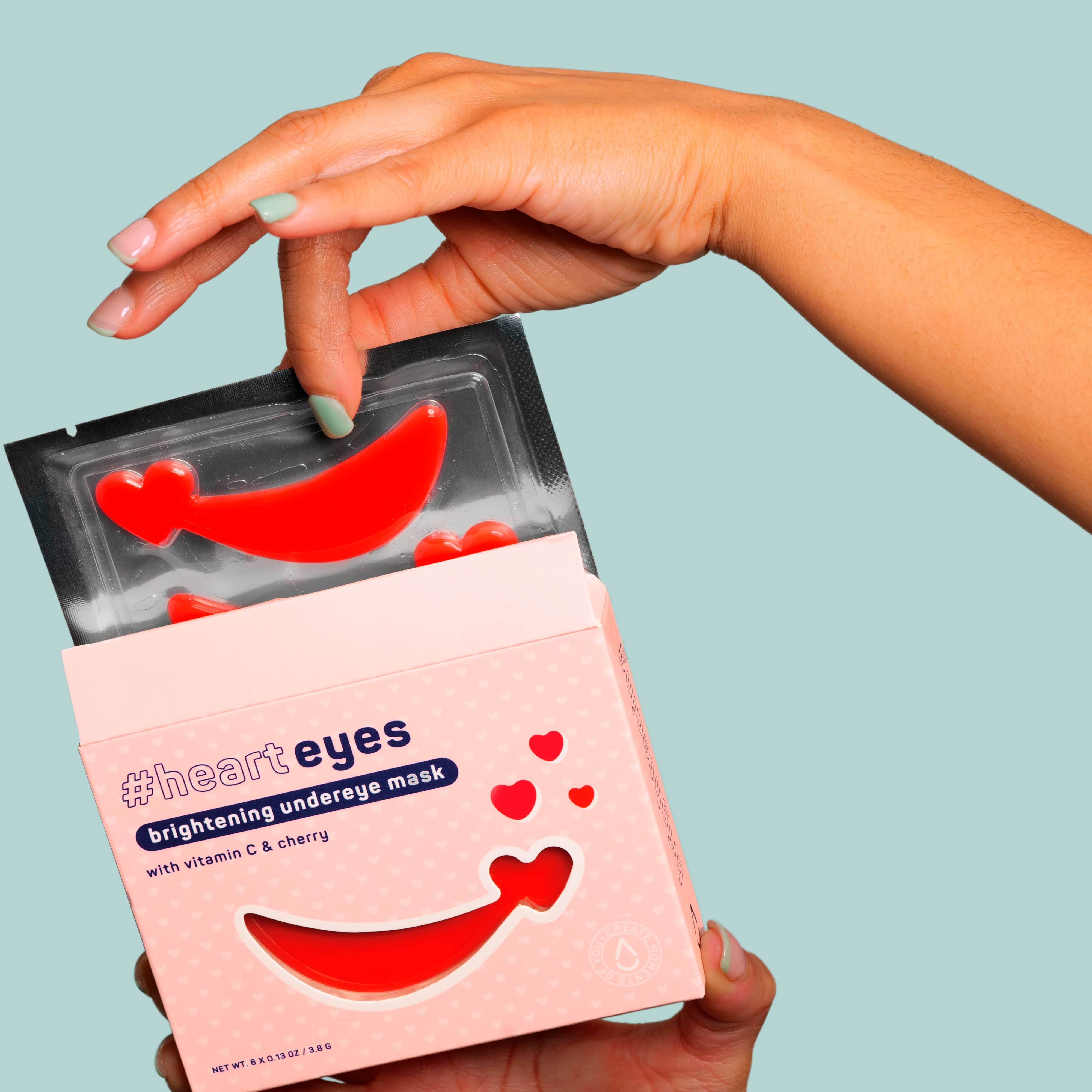 A hand with pastel green nails holds a red under-eye mask above an open pink box labeled "#heart eyes." The mood is fresh and playful.