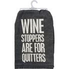 Dark gray towel draped over a hanger, featuring bold white text: "WINE STOPPERS ARE FOR QUITTERS." Humorous tone, kitchen decor.
