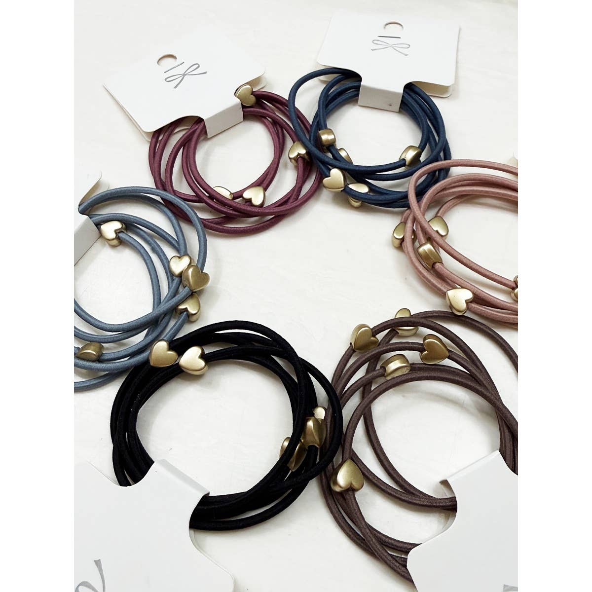 Six sets of hair ties in various colors—burgundy, blue, pink, black, gray, and brown—each adorned with small gold heart charms, are arranged on a white surface.