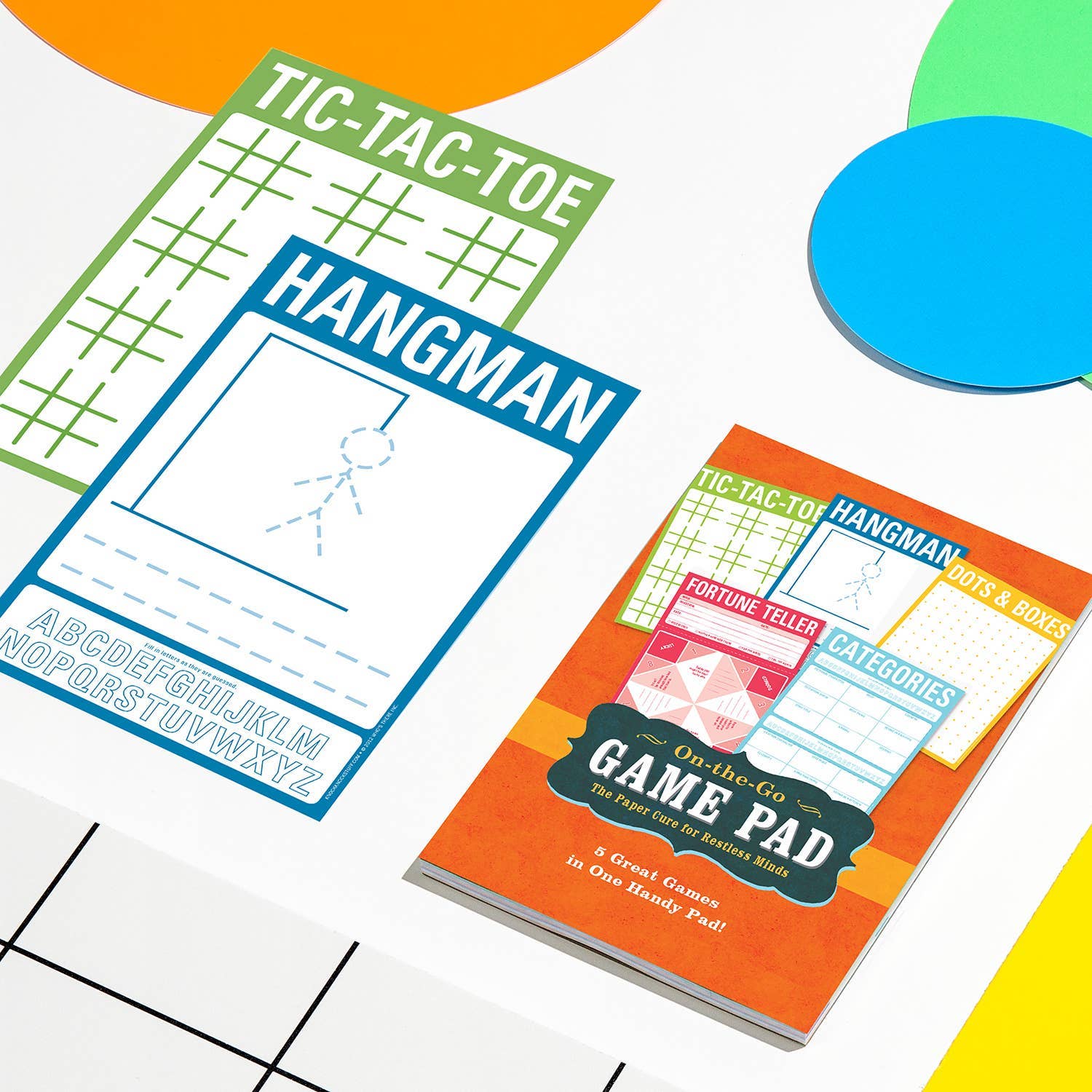 Colorful activity pads featuring tic-tac-toe and hangman designs on a bright table with circular, colorful shapes. Playful and engaging tone.