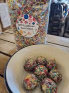 Bowl of colorful seed bombs on a wooden table. Behind them, a bag labeled "DIY Wildflower Seed Bombs" filled with multicolored shredded material.