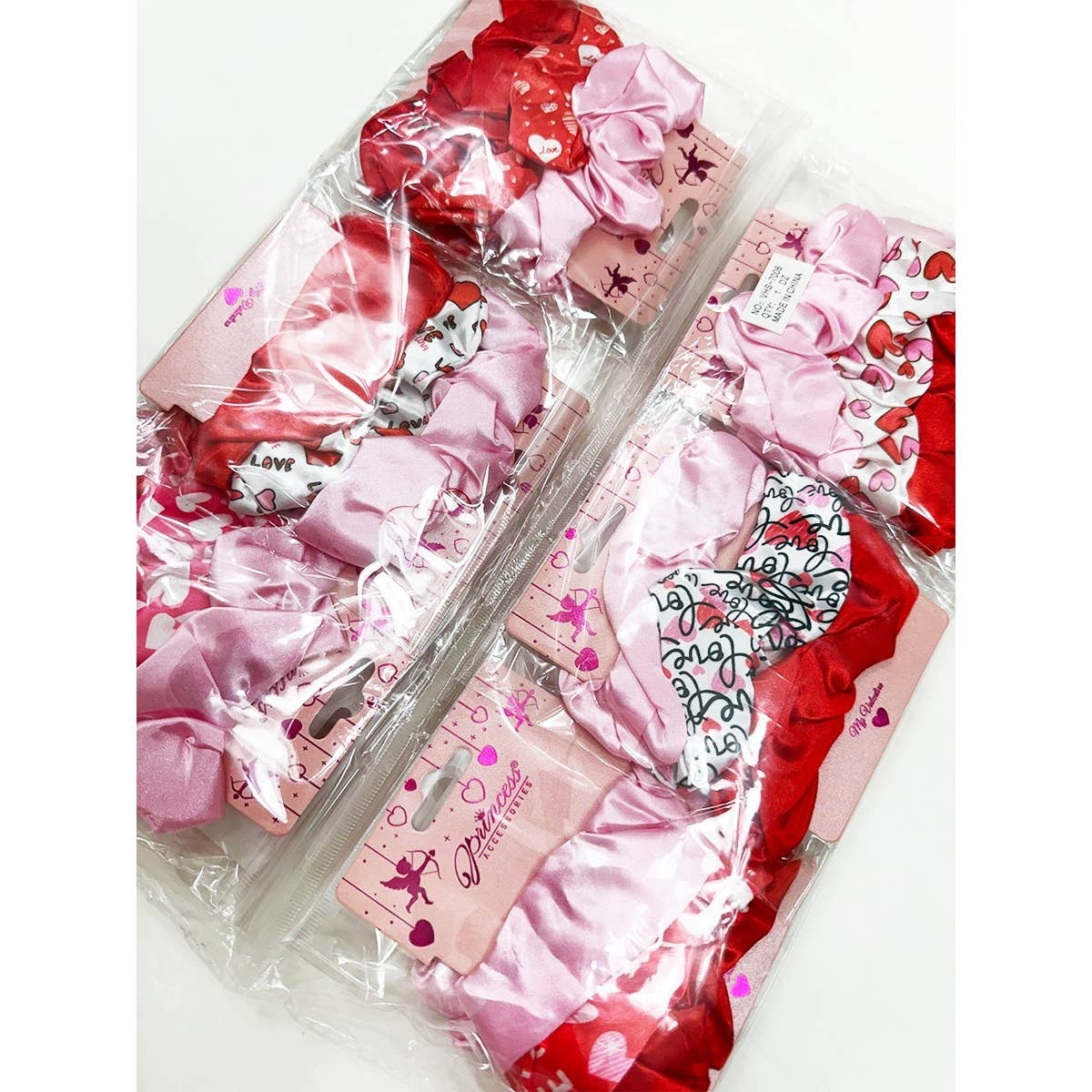Packaged sets of hair scrunchies in pink and red with heart patterns. Labels read "Princess Accessories" for a cute, playful style.