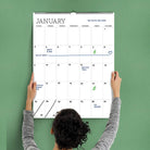 A person hangs a large January calendar on a green wall. Key dates include a family trip, dad's birthday, a dentist appointment, and an oil change.