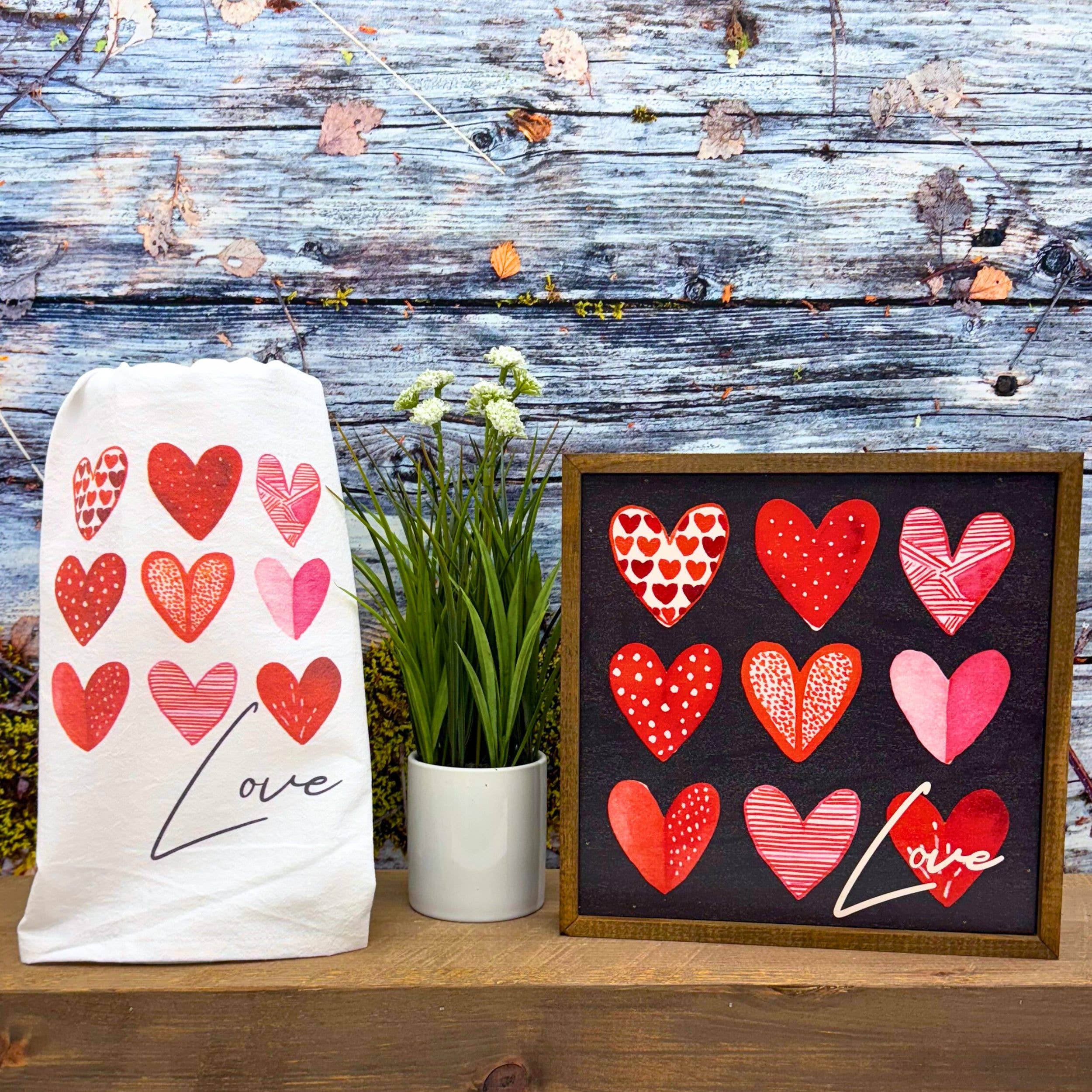 Decorative towel and frame with red and pink heart patterns, with the word "Love" scripted in black and white. A small potted plant sits between them, against a rustic wooden backdrop.