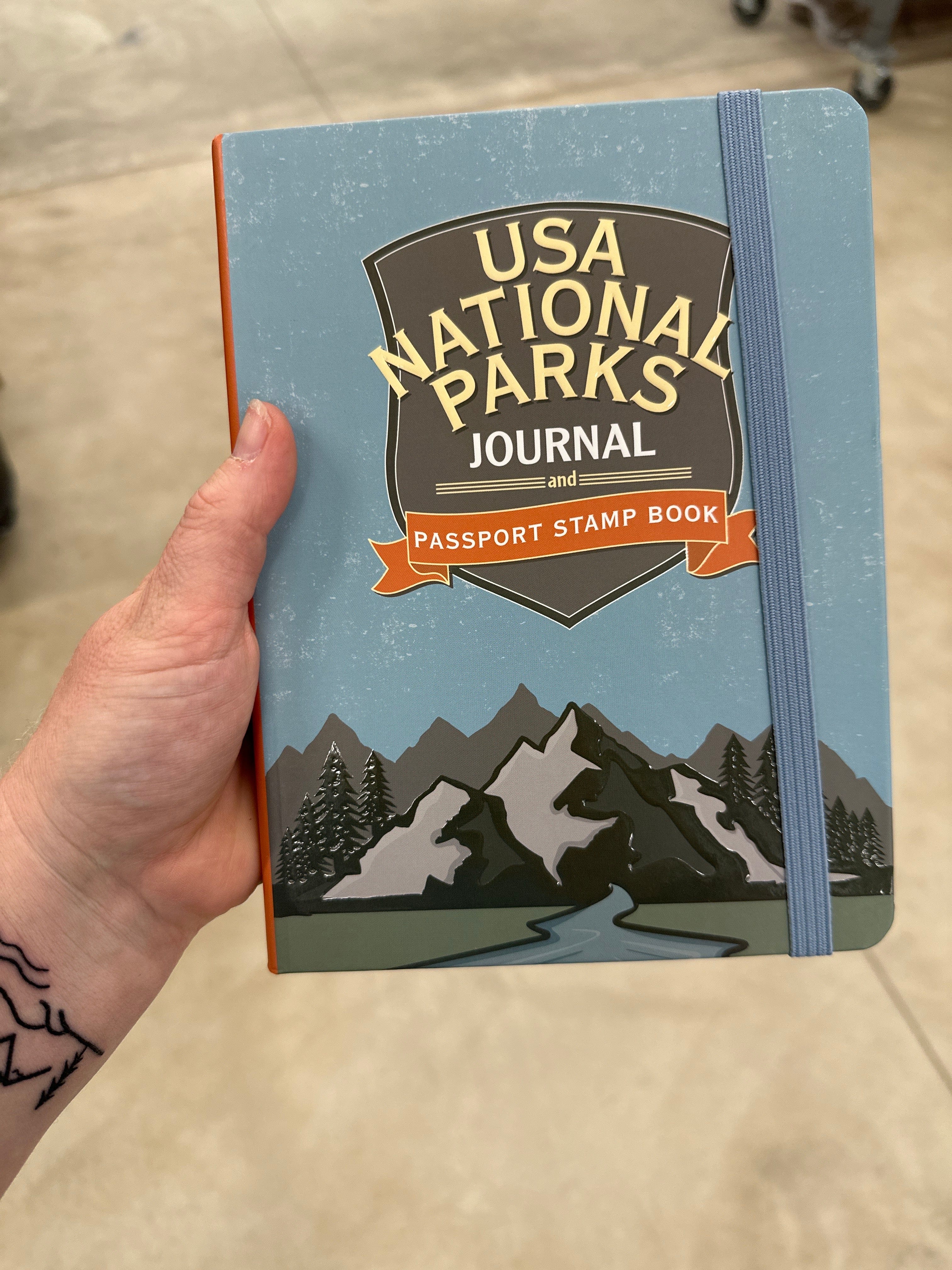 Person holding a "USA National Parks Journal and Passport Stamp Book" with mountain illustration on the cover. The tone is adventurous and inviting.