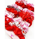 A collection of vibrant scrunchies in red and pink hues, some patterned with hearts and words, displayed on love-themed packaging with a playful, romantic vibe.