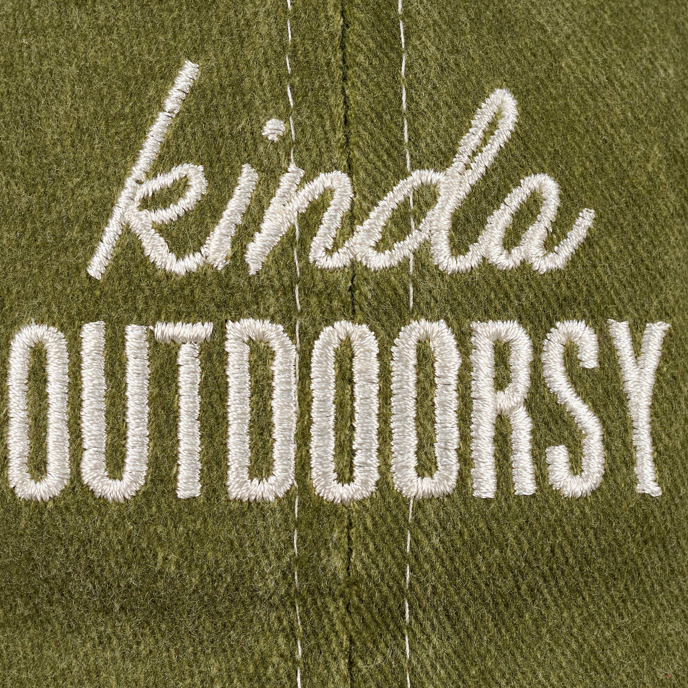 Close-up of green fabric with white embroidered text reading "kinda outdoorsy." The playful font and earthy tone suggest a casual, nature-themed vibe.