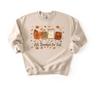 A beige crewneck sweatshirt featuring a cute autumn-themed graphic of three books with faces, surrounded by falling leaves and acorns, with the text 'All Booked for Fall' printed below.