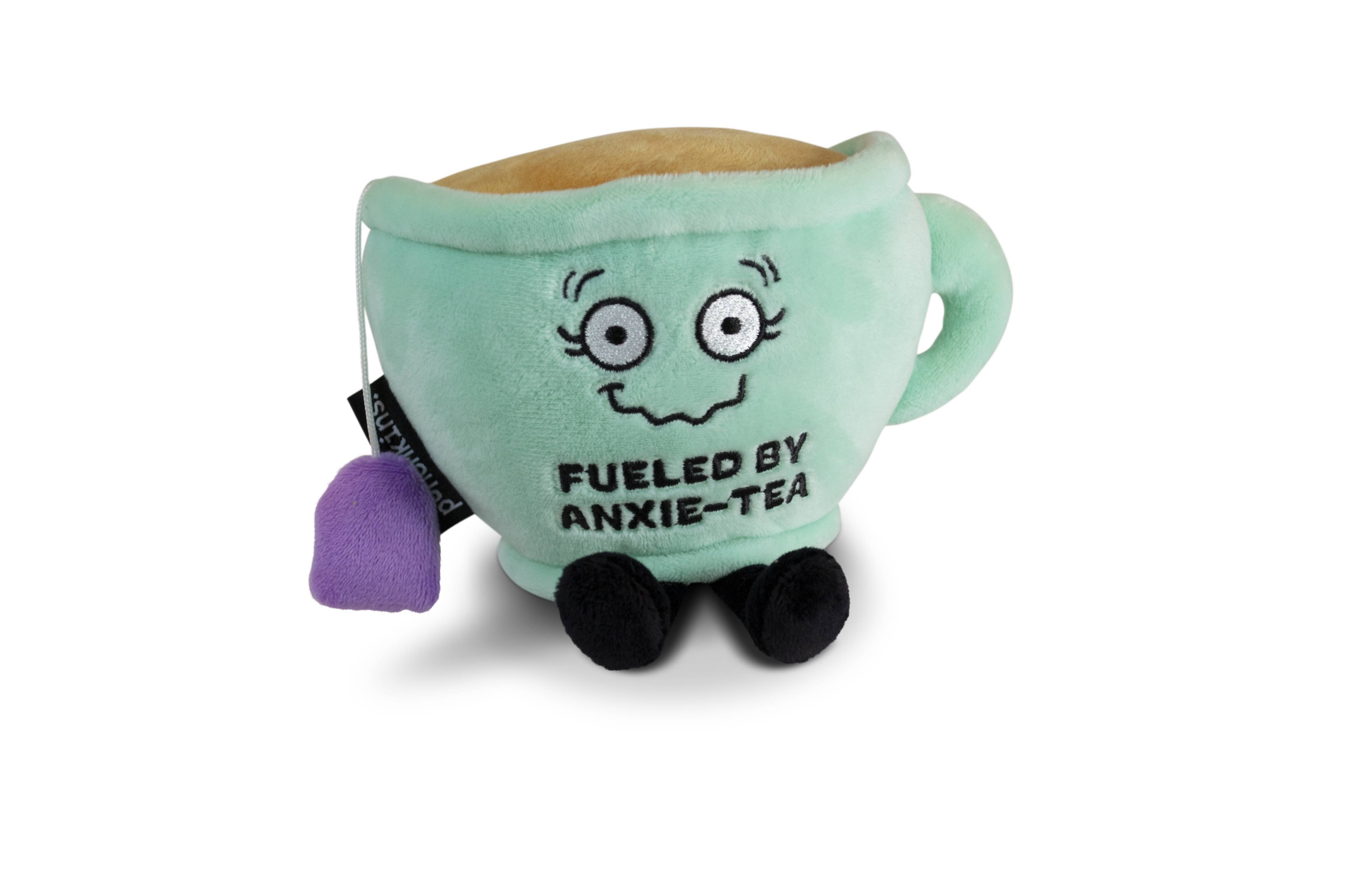 A green plush toy shaped like a smiling tea cup with a shaky smile. The cup is filled with soft, plush 'tea' on top, and a purple tea bag hangs from the side. The front displays the embroidered phrase "FUELED BY ANXIE-TEA" in bold letters. It has small black legs, a handle on one side, and an overall cute, cozy design.