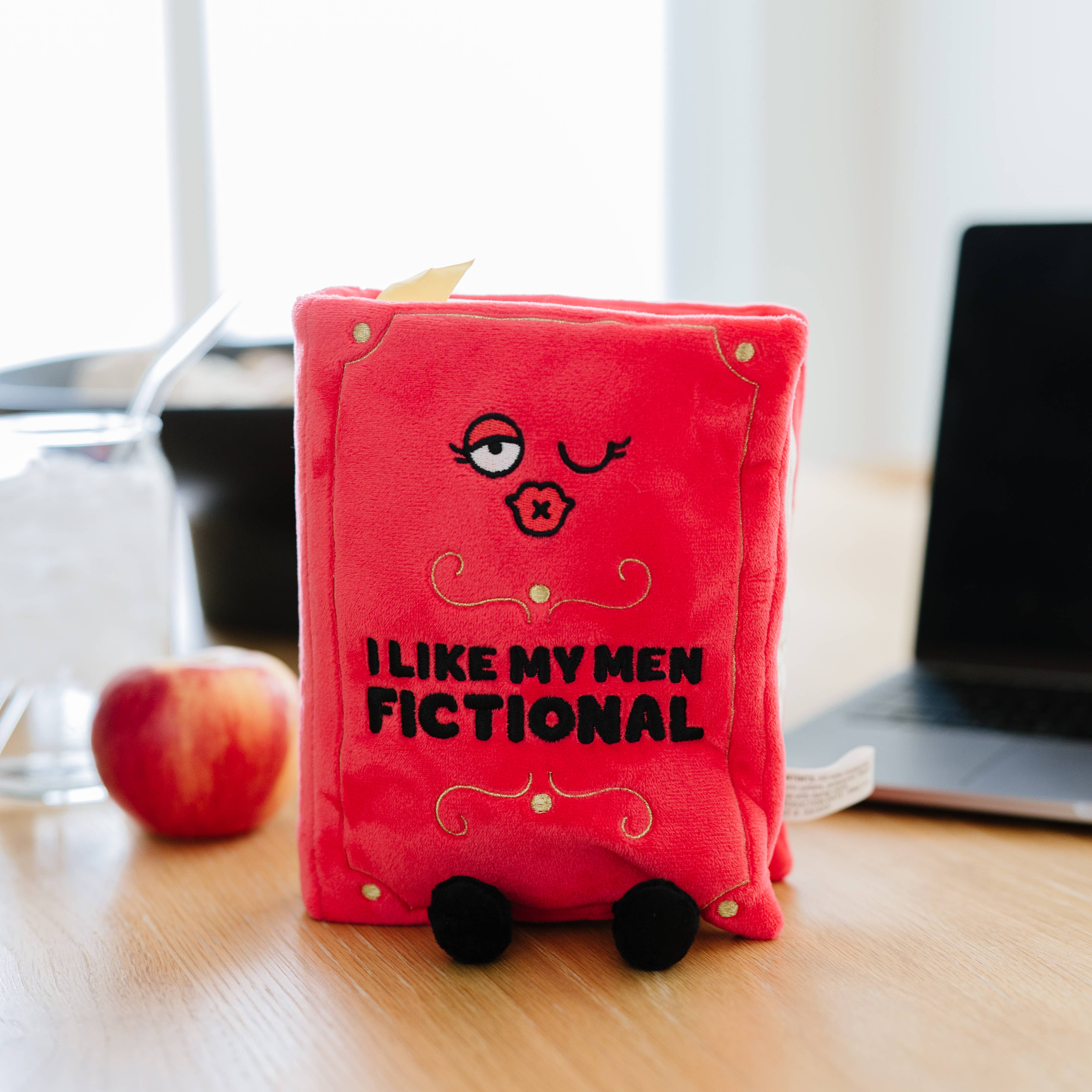 A red plush book with a kissy winky face and embroidered black text that reads 'I like my men Fictional.' The plush has black legs sticking out, a yellow bookmark peeking from the top, and 'A Spicy Romance' embroidered in black on the book's spine. The plush is sitting on a desk in front of a laptop and apple.