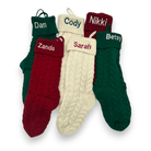 Four personalized cable-knit Christmas socks in green, cream, and burgundy, arranged together on a white background. Each sock has a different name embroidered on the cuff: Dan, Cody, Nikki, and Betsy, with Zanda also visible on a burgundy sock.