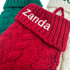 A close-up of a red cable-knit sock holder with a folded cuff embroidered with the name 'Zanda' in white. Other knitted holders in green and cream are partially visible in the background.