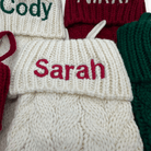 Close-up of a cream knit Christmas stocking with the name 'Sarah' embroidered in red across the cuff, surrounded by other knitted stockings in red, green, and cream.