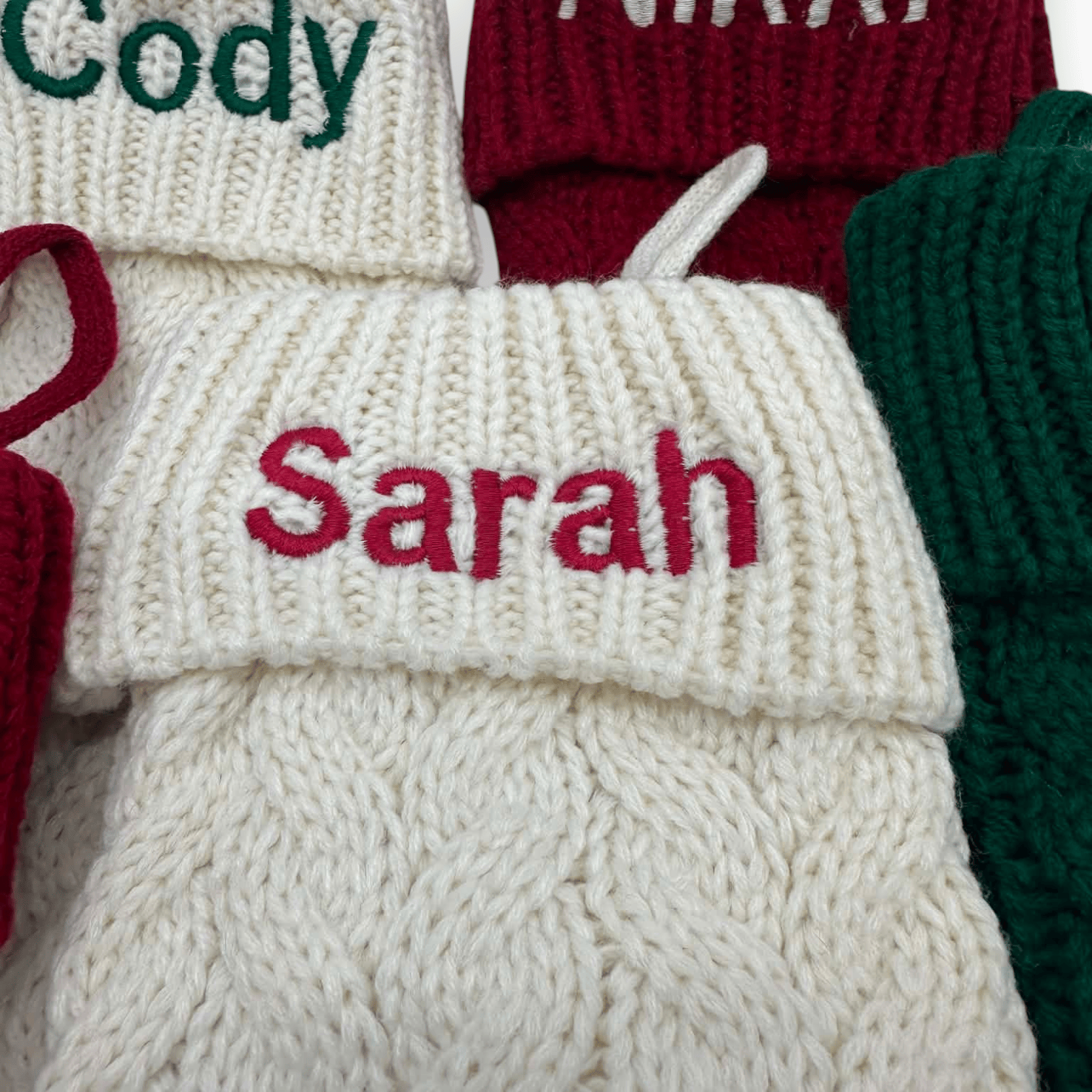 Close-up of a cream knit Christmas stocking with the name 'Sarah' embroidered in red across the cuff, surrounded by other knitted stockings in red, green, and cream.