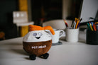 A brown and white coffee pot plushie with an orange pour spout. The white portion has a smiley face, and the brown portion features white text that says "Pot Head." The plushie has black legs sticking out. Plushie is sitting on a desk