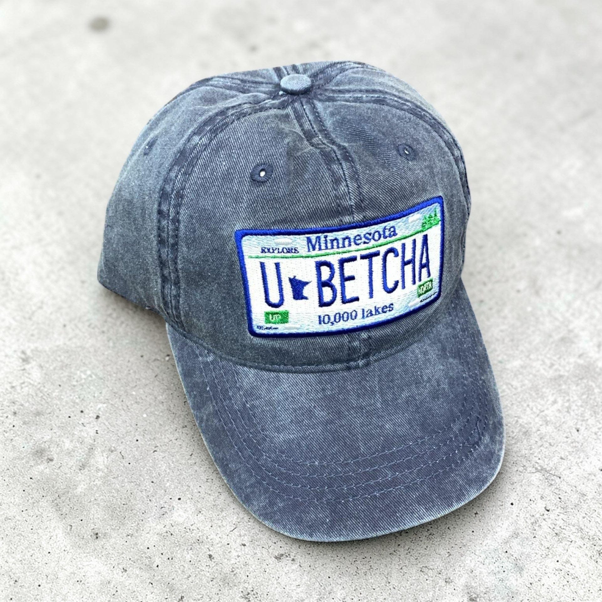 Blue cap with a Minnesota-themed license plate design that states “u betcha” on a concrete surface. 