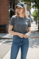 Woman wearing a gray t-shirt and blue jeans and Uff Da embroidered hat standing on a sidewalk.