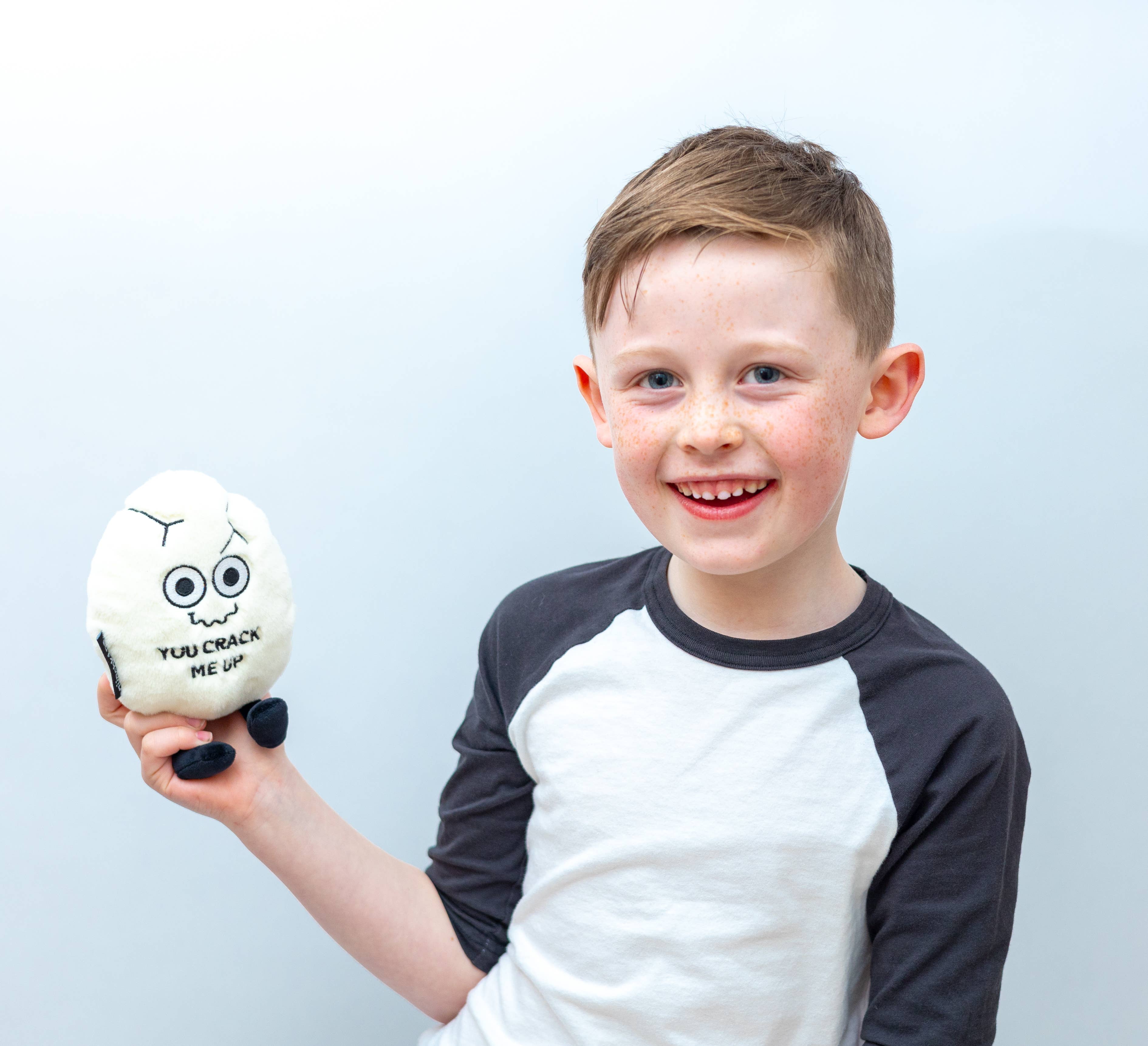 Child holding a white cracked egg plush with a squiggly smiley face and black text that says "You crack me up." The plushie has black legs sticking out.