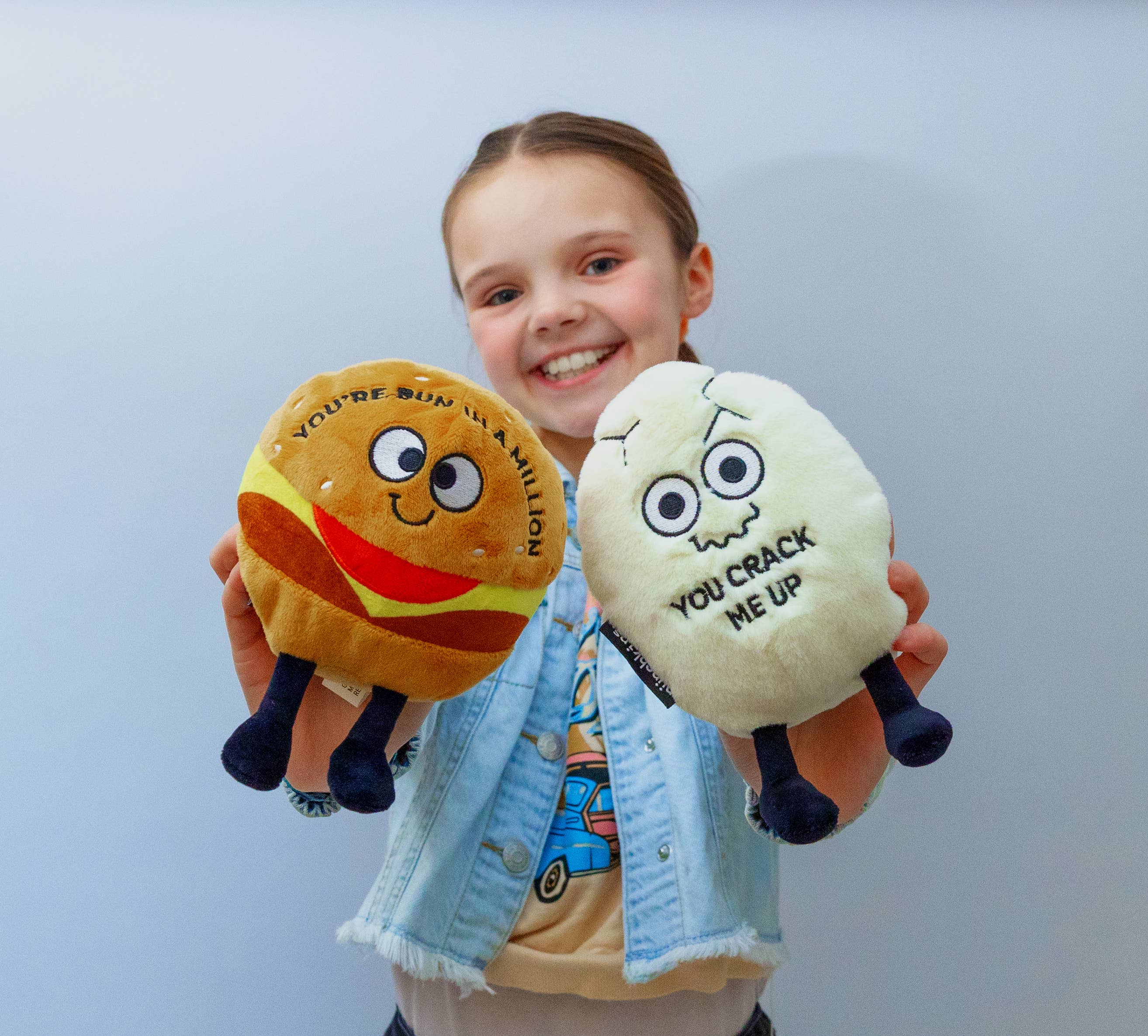 Child holding two plushies. Awhite cracked egg plush with a squiggly smiley face and black text that says "You crack me up." The plushie has black legs sticking out. The other a hamburger plushie