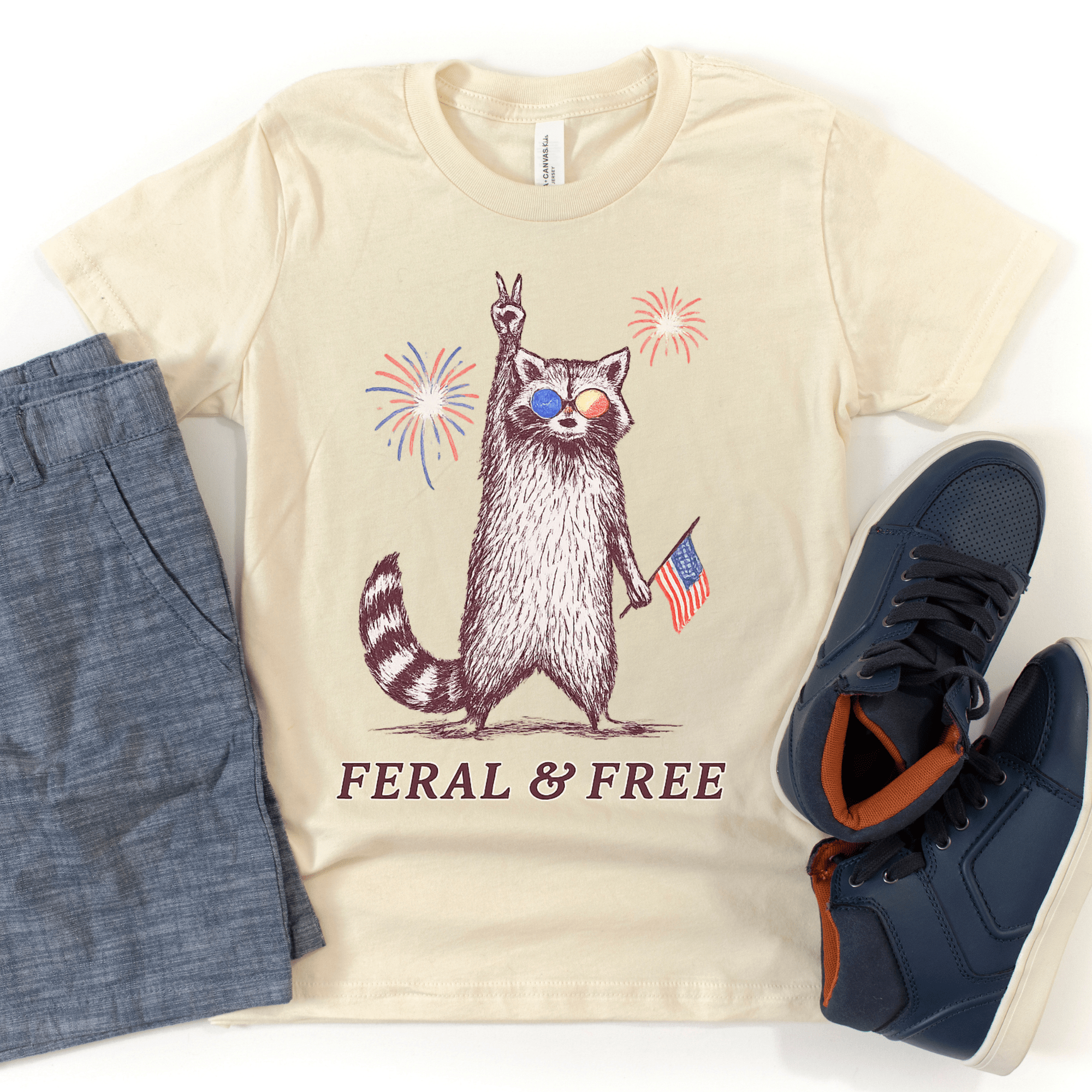 Beige t-shirt with a raccoon graphic and text, paired with blue shorts and navy sneakers on a white background.