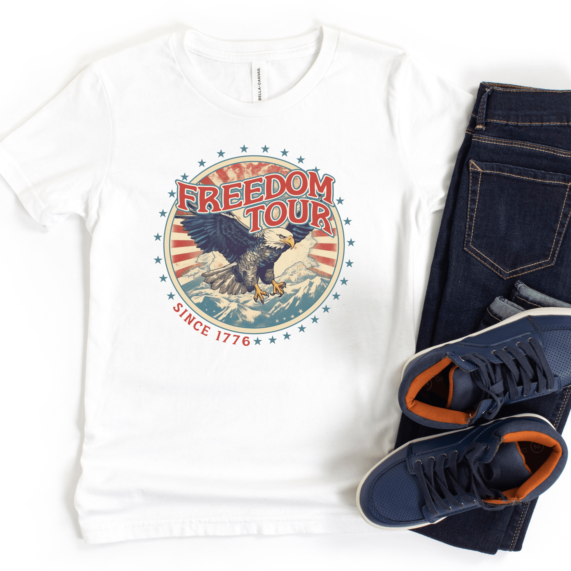 A white kids' t-shirt with a patriotic 'Freedom Tour Since 1776' print, displayed alongside blue jeans and a pair of sneakers.