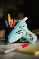 A blue glue gun plushie with a smiley face, three flowers, and black embroidered text that says 'I'm a crafty bitch.