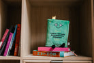 A green plush with a smily face and embroidered black text that reads 'I like fantasies with impressive wingspans.' The plush has black legs sticking out and a yellow bookmark peeking from the top. Plush is sitting on a bockshelf.