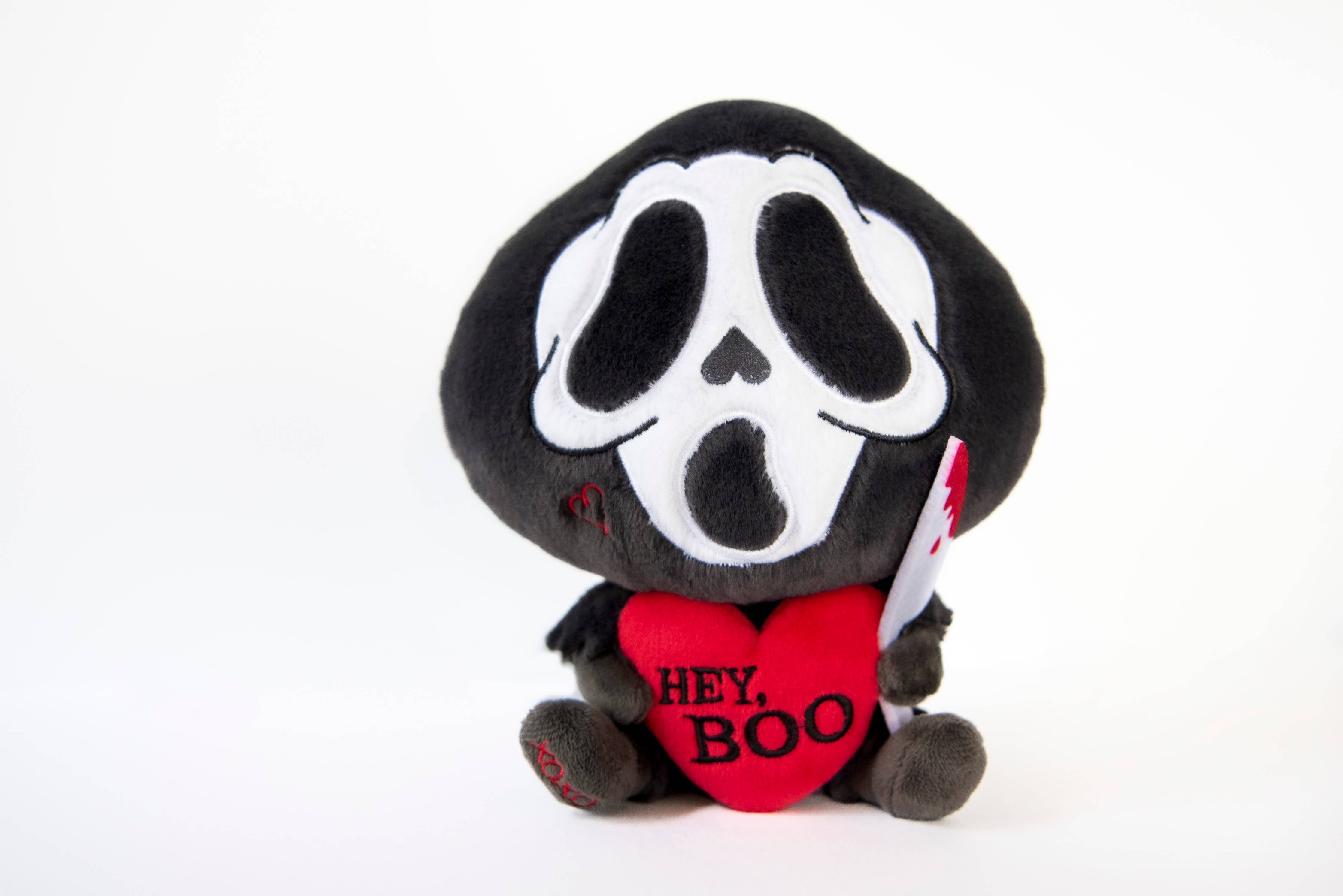 A black and white ghost resembling the Scream mask, holding a bloody knife in one hand and a red heart in the other. The heart has the phrase "Hey, Boo" written in black text.