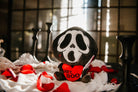 A black and white ghost resembling the Scream mask, holding a bloody knife in one hand and a red heart in the other. The heart has the phrase "Hey, Boo" written in black text.