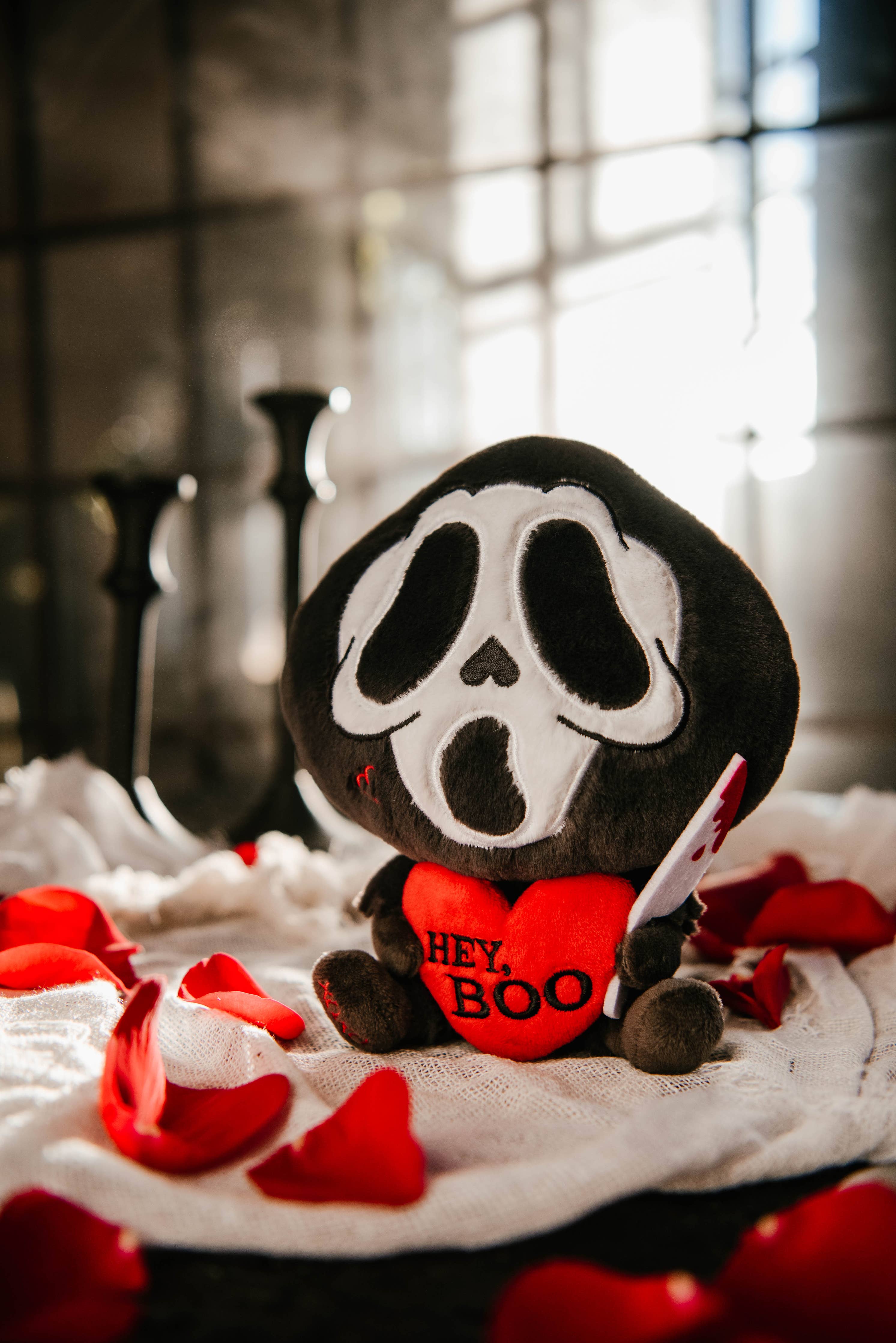 A black and white ghost resembling the Scream mask, holding a bloody knife in one hand and a red heart in the other. The heart has the phrase "Hey, Boo" written in black text.