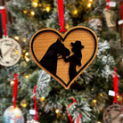 A wooden ornament shaped like a heart with a silhouette of a horse and a girl within the heart, hanging from a Christmas tree with other ornaments.