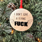 A wooden ornament with the phrase 'I DON'T GIVE A FLYING FUCK' cut out in the center, mounted on a red ribbon, against a backdrop of a Christmas tree.