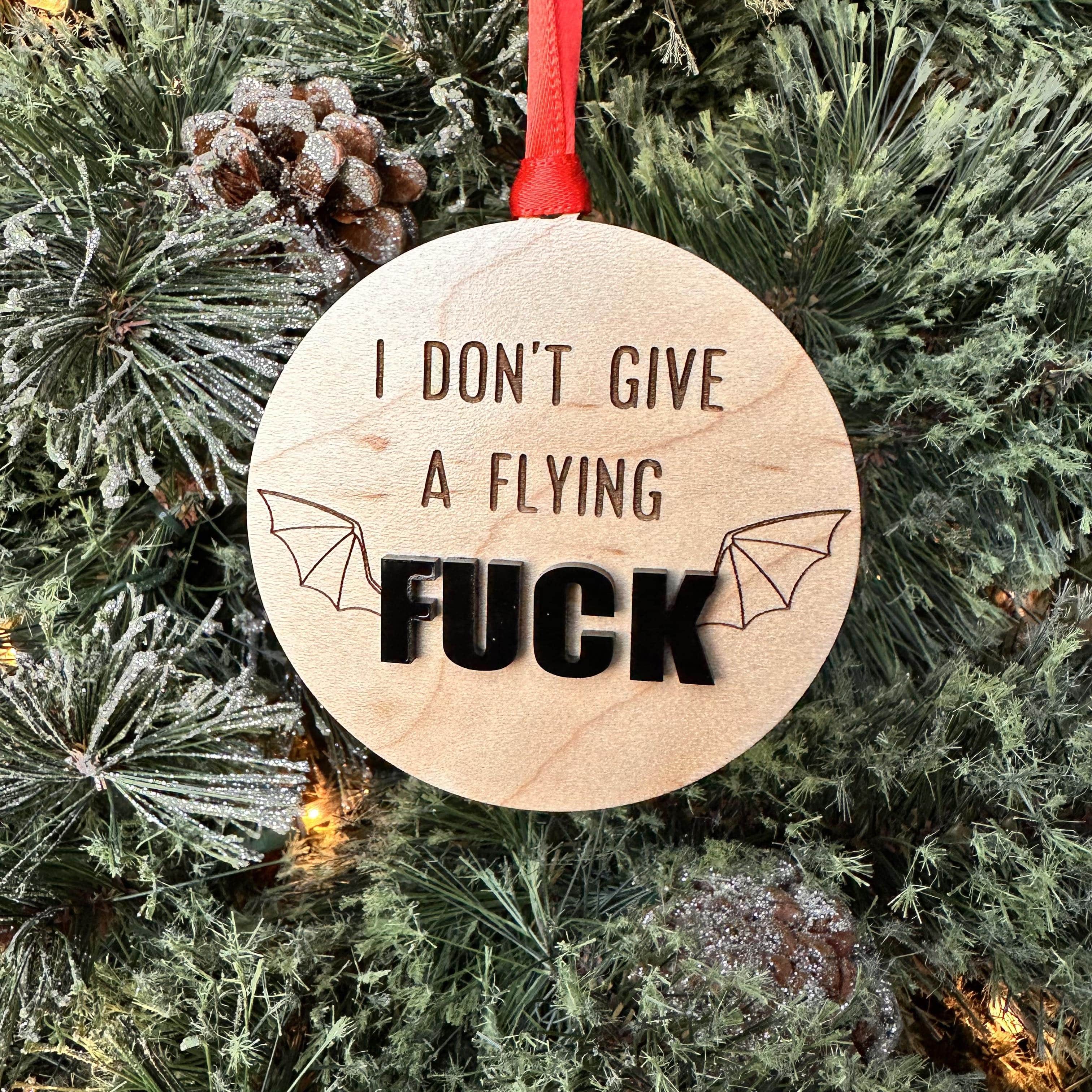 A wooden ornament with the phrase 'I DON'T GIVE A FLYING FUCK' cut out in the center, mounted on a red ribbon, against a backdrop of a Christmas tree.