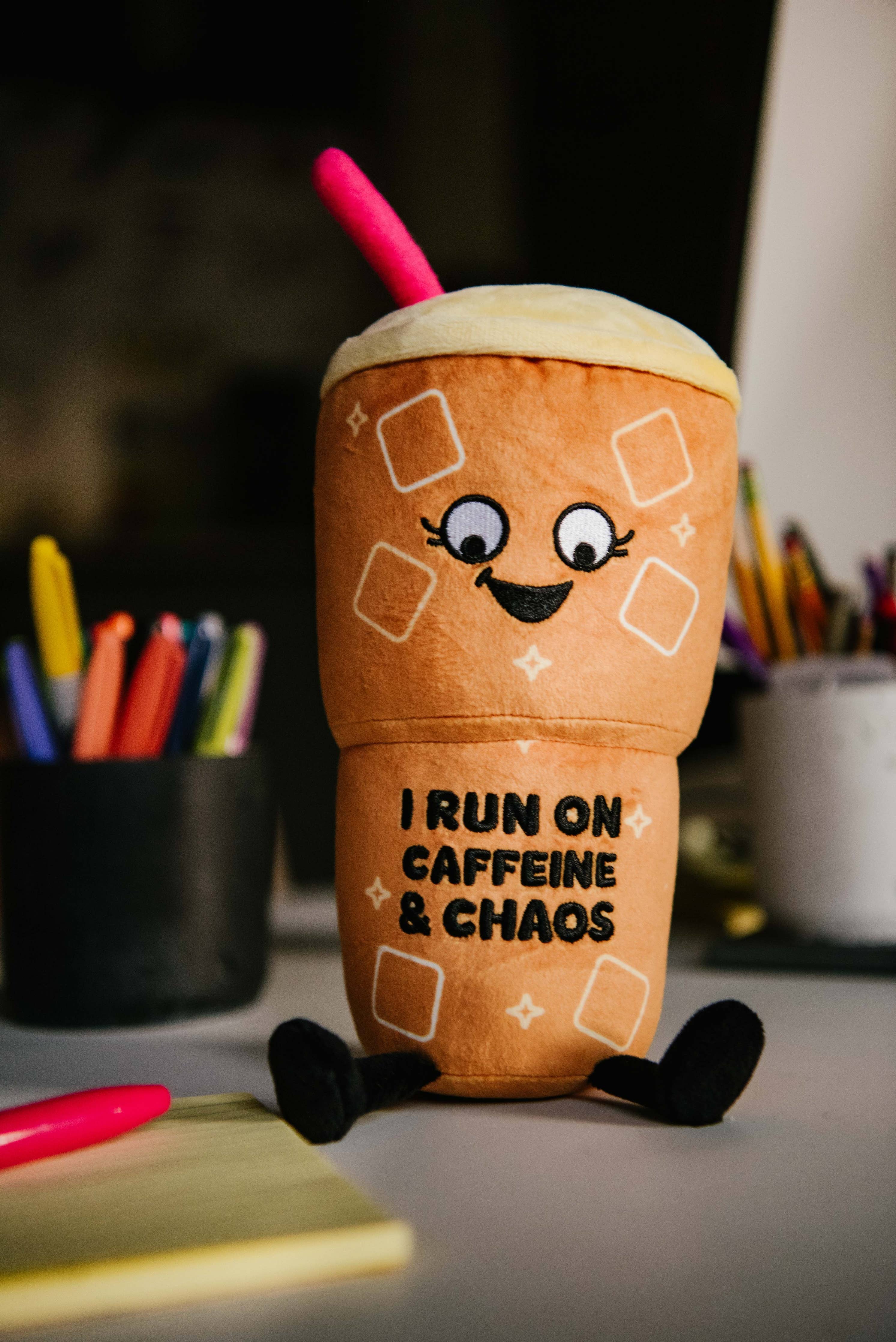 Plushie shaped like a ln iced coffee with a cute face that reads “ I run on caffeine and chaos”