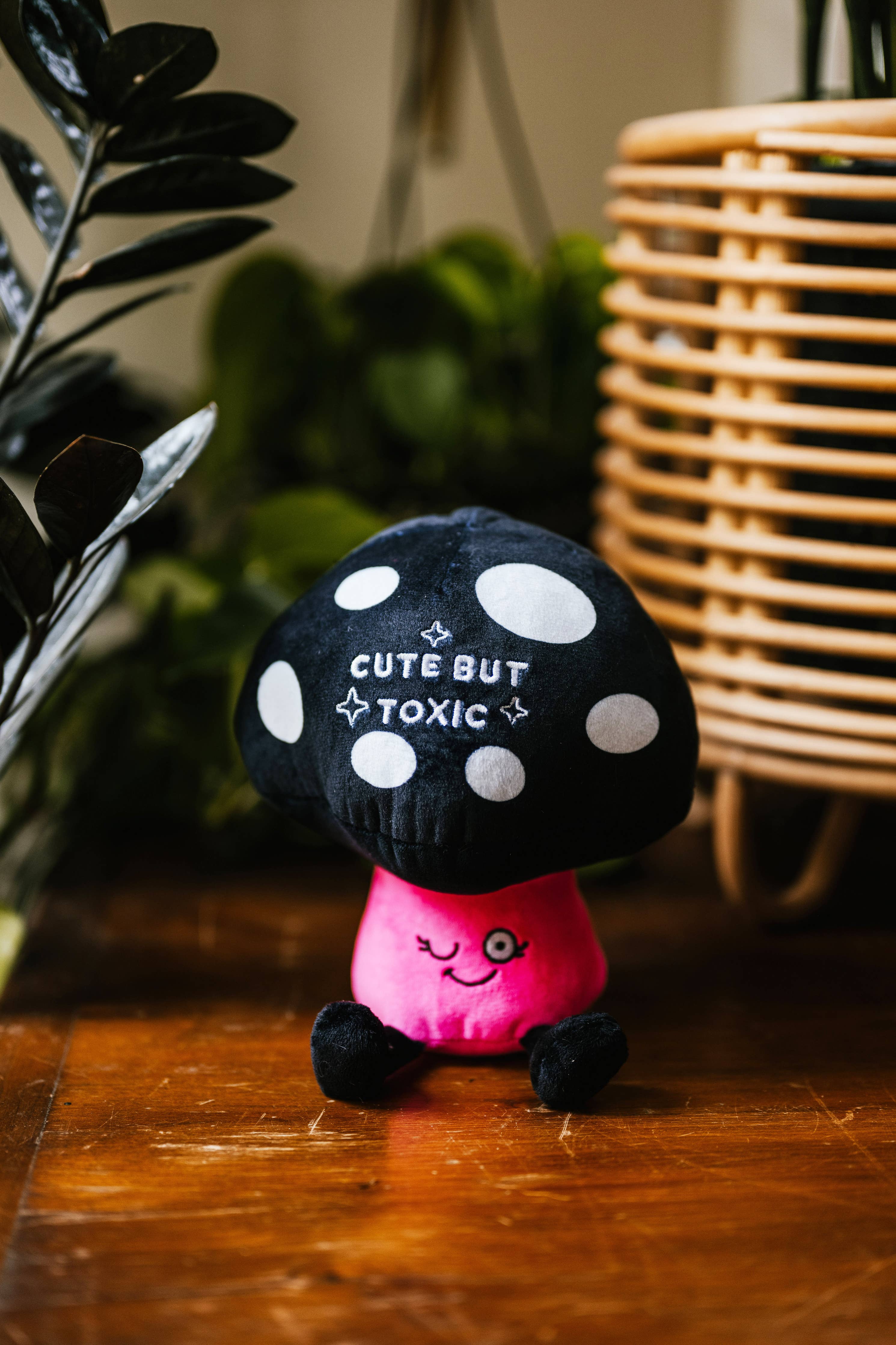 A mushroom plush with a black and white polka dot top and white text that says "Cute but toxic." The stem is pink with a winking smiley face, and the plushie has black legs sticking out.