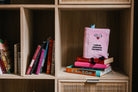 Light pink plush book sitting up with kissy face and black embroidered words that say 'I like my romances historical.' Black legs sticking out bottom and a blue bookmark peeking out the top.  Plush is sitting on a bookshelf.