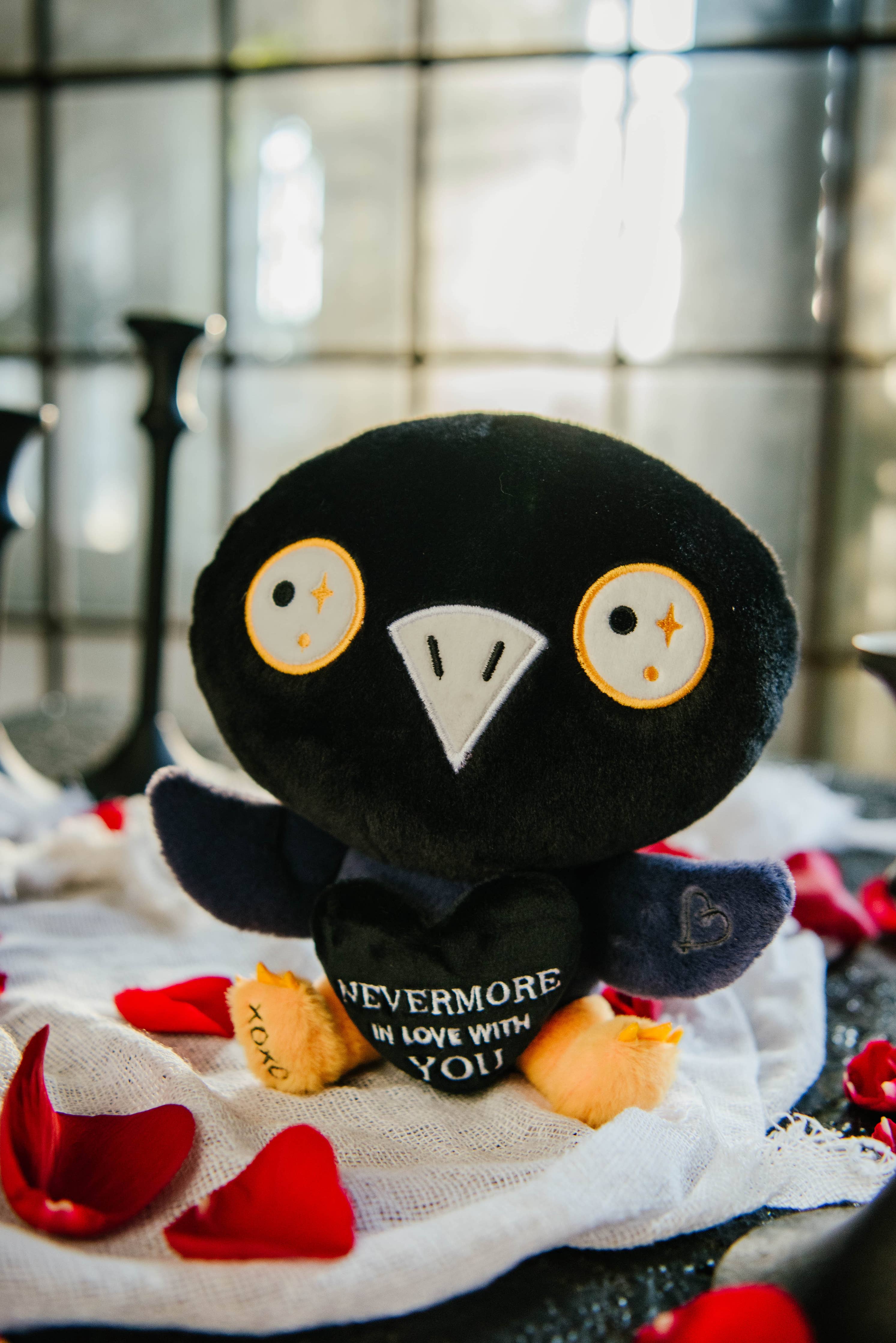 A black and blue raven plushie with a white beak and white and yellow eyes. Its wings are holding a black heart with the white text "Nevermore in love with you." The raven has yellow legs with a black "xoxo" on the right foot.